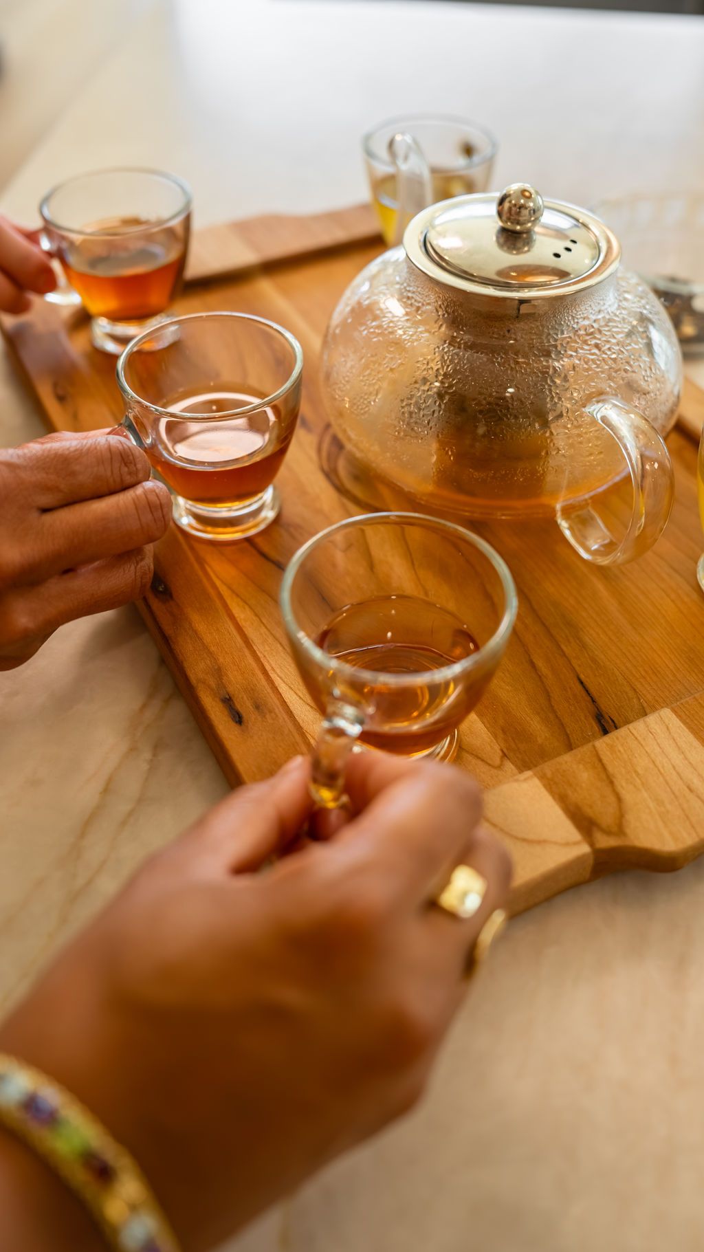 A glass teapot and small glasses of golden tea on a wooden tray, with hands reaching to share a cup